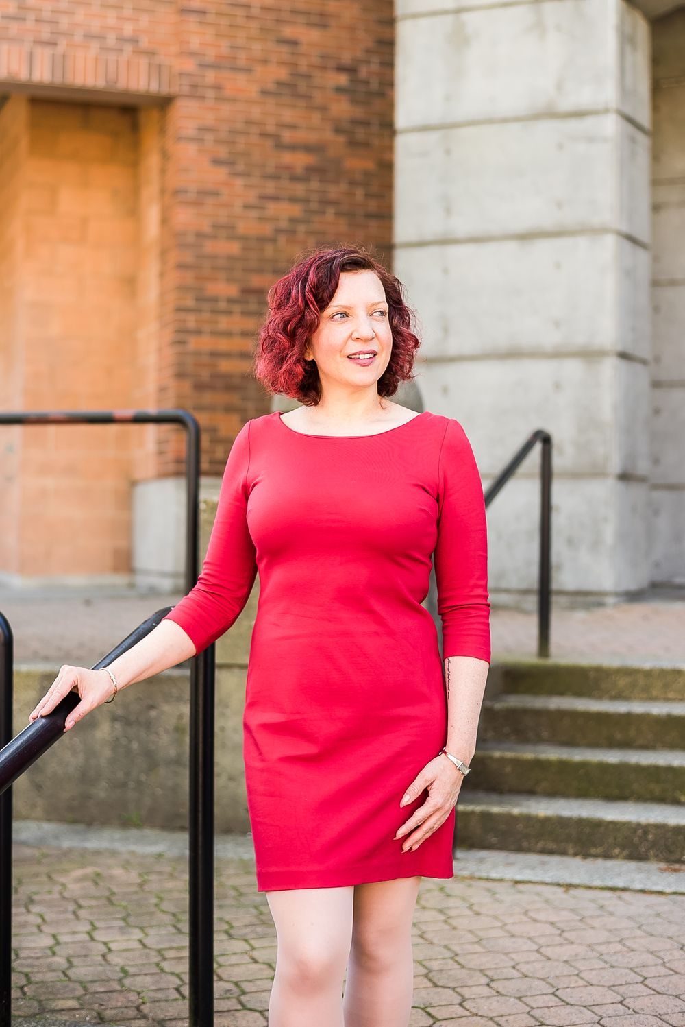 A woman wearing a red dress poses outside on a staircase with a brick wall in the background.