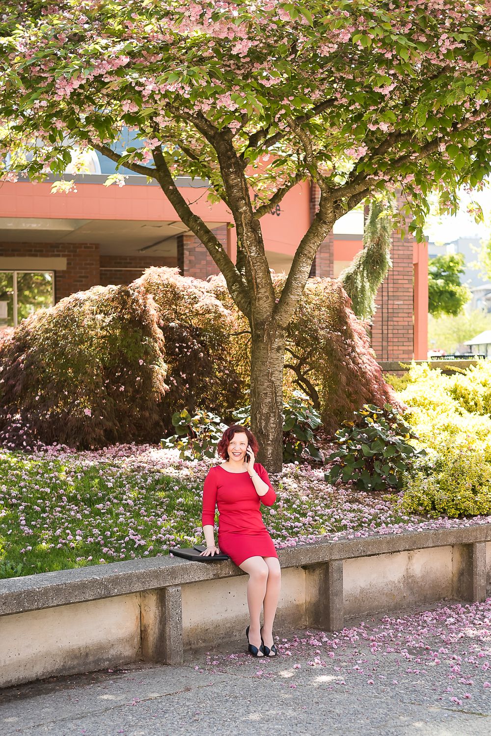Under a cherry blossom tree in the spring, a woman wearing a bright red dress takes a phone call for work.