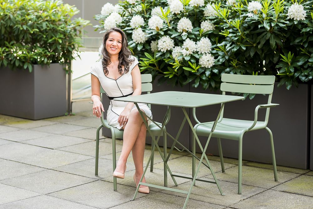 A businesswoman dressed in a white dress sits casually at an outdoor patio table with flowers behind her.