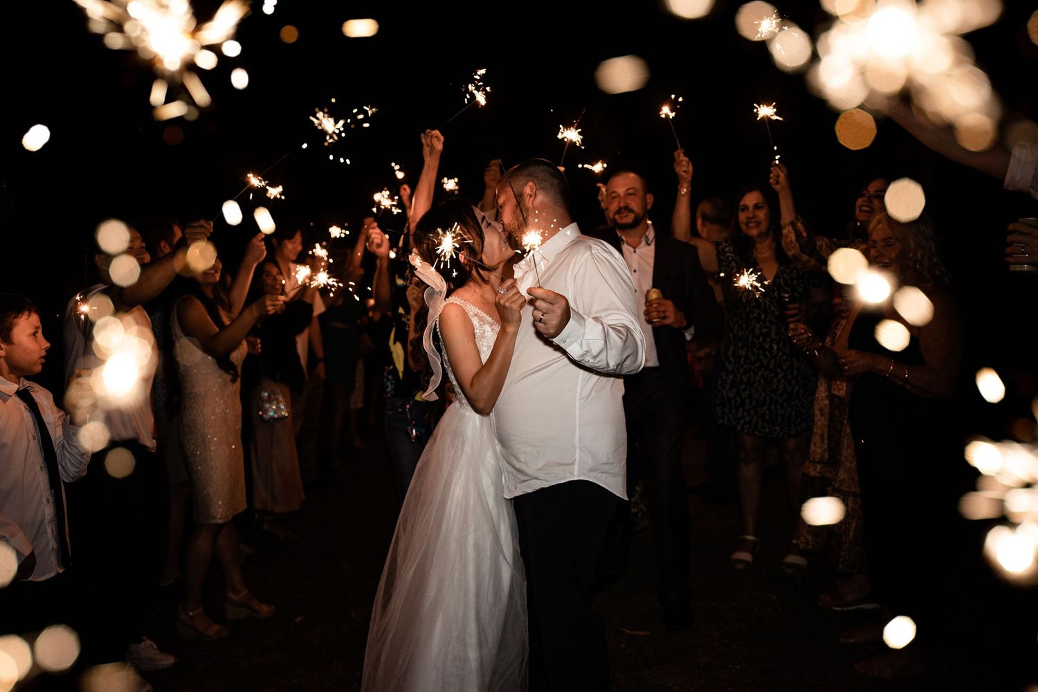 Wedding couple dances surrounded by sparkler lights and celebrating guests at night.