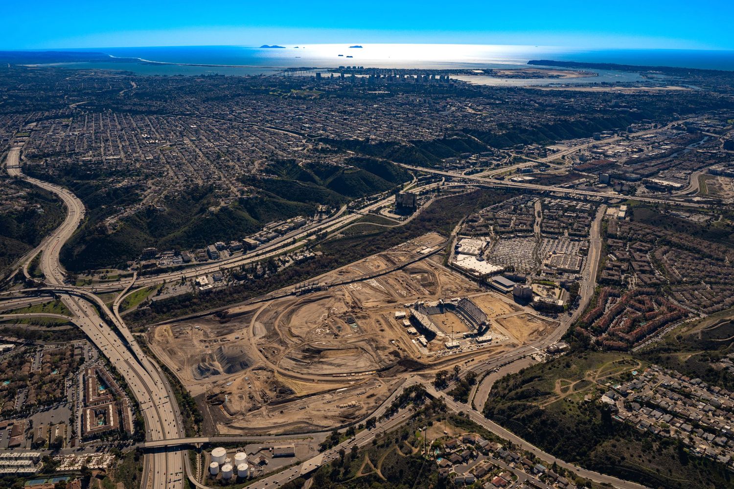 High-altitude aerial shot showing extensive earthwork, site grading, and early stadium framing at a large-scale urban development project.