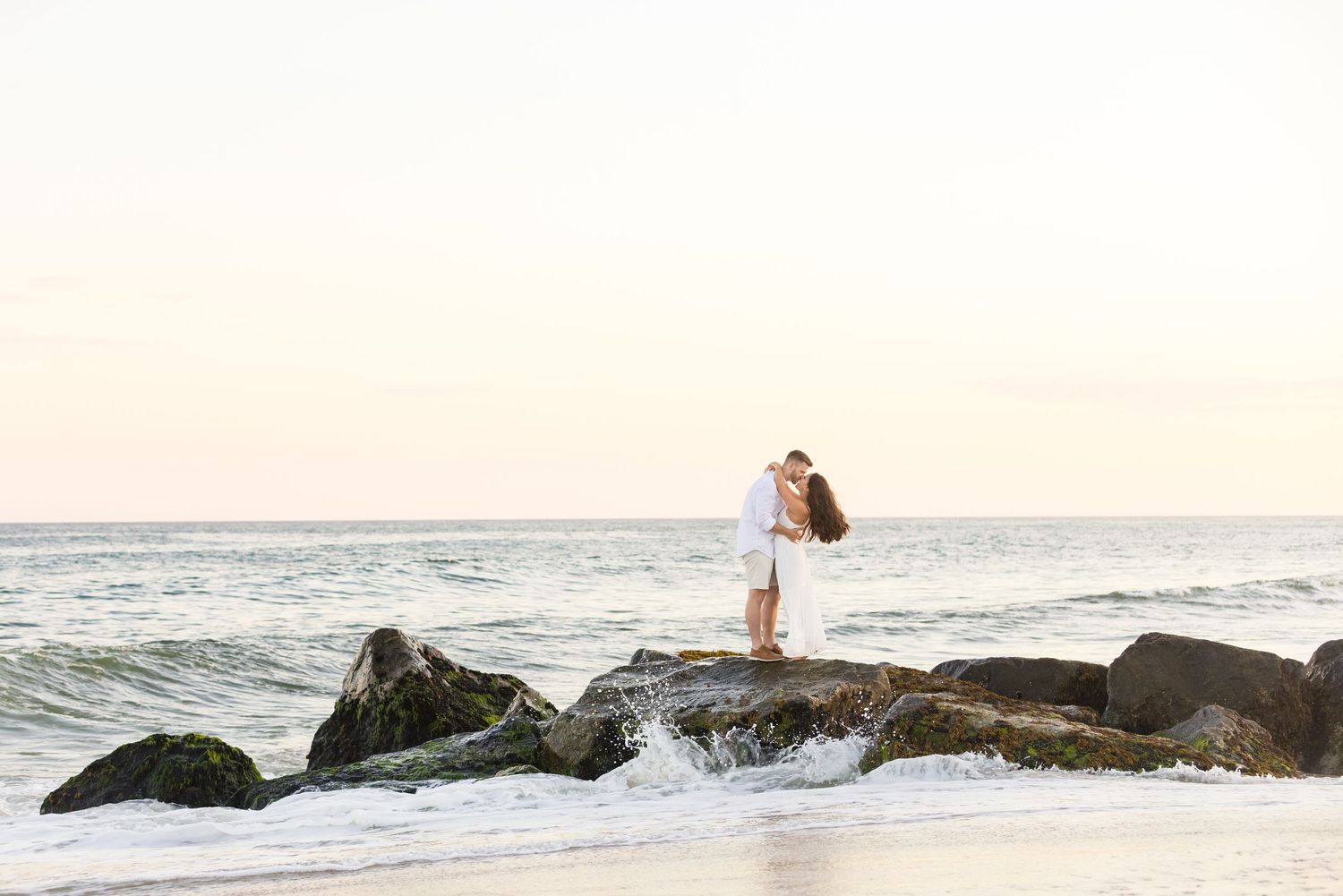 Romantic sunset beach photo with waves crashing against rocks along the shoreline.