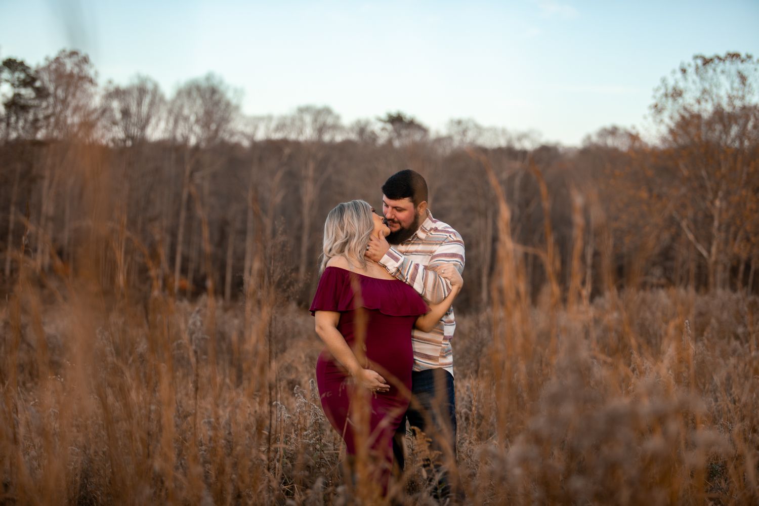 A couple shares a romantic moment in a field of dried grass during sunset.