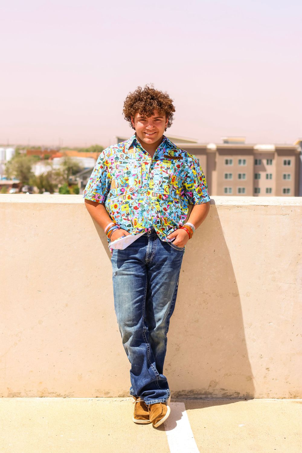 Young child in colorful floral shirt and jeans sitting casually on concrete wall outdoors.