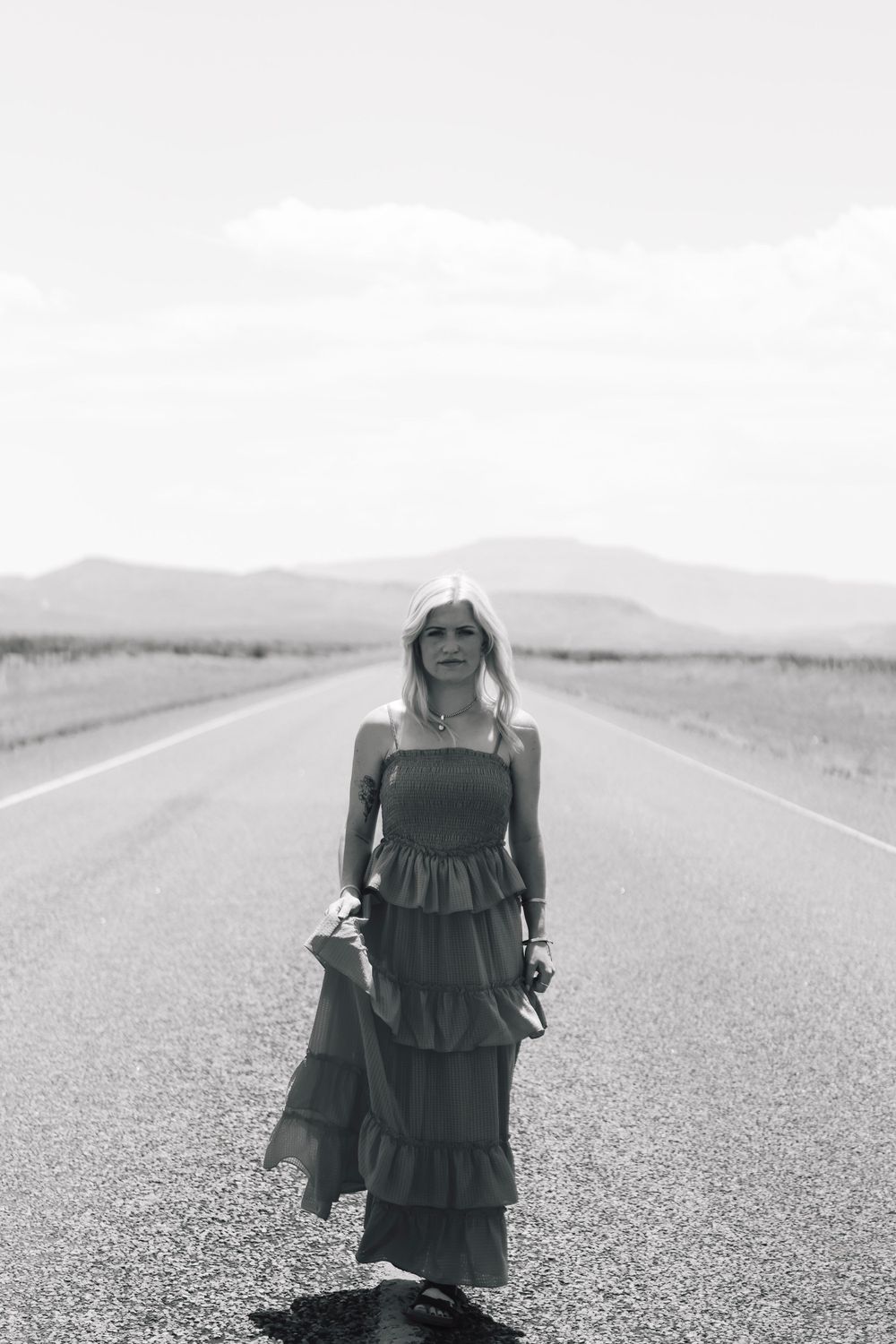 A black and white photo of a figure in a tiered maxi dress standing on an empty desert road.
