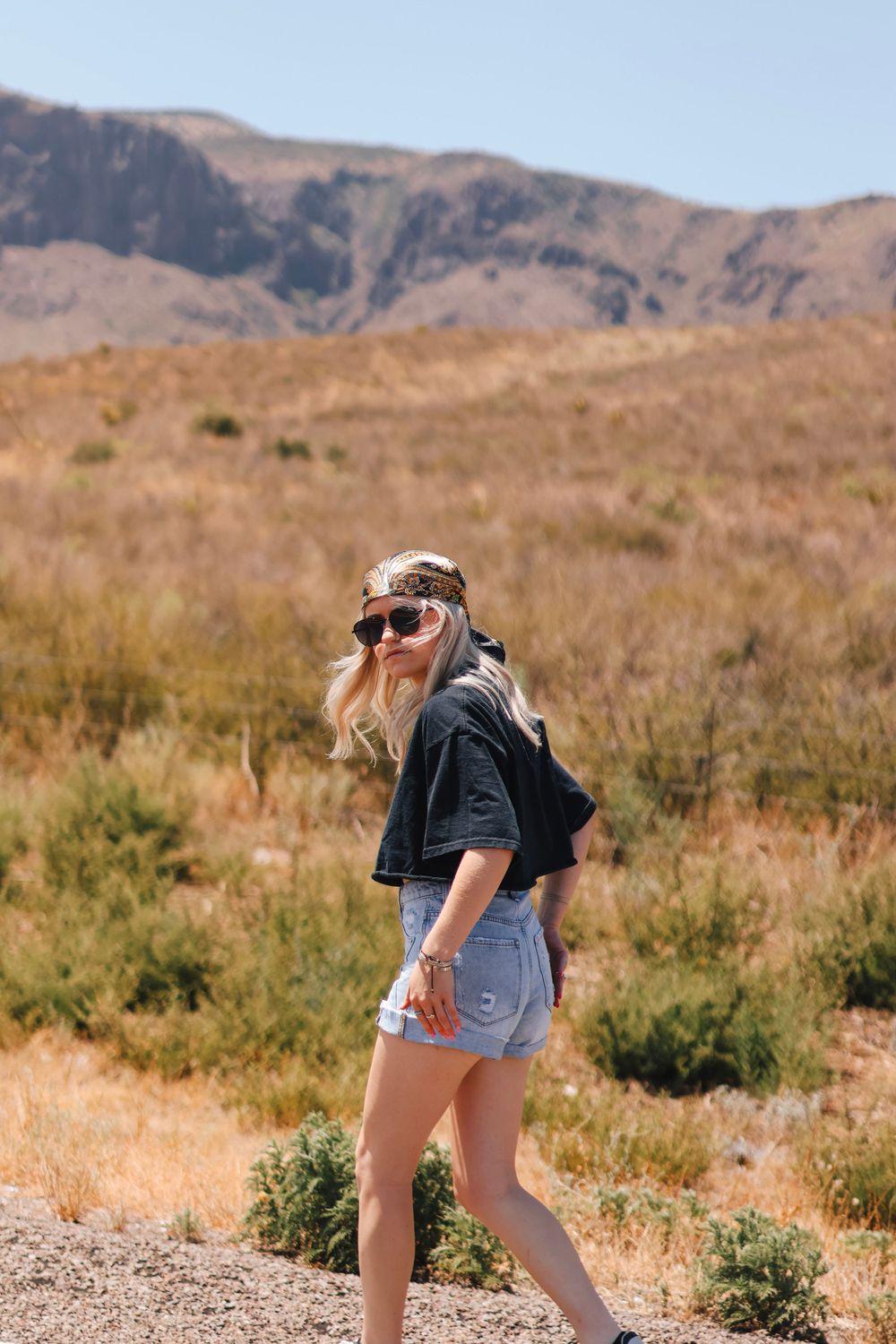A person in denim shorts and jacket poses against a desert mountain landscape.