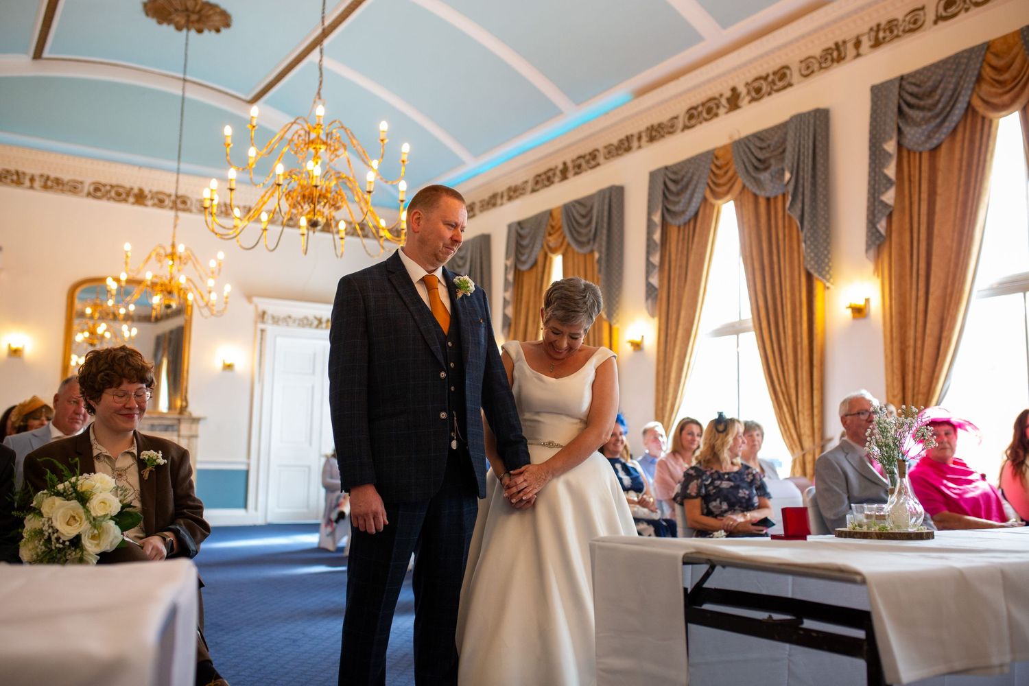 Wedding couple holding hands during ceremony in ornate room with chandeliers and gold drapes.