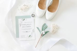 White wedding shoes, stationery, and greenery on a marble surface.