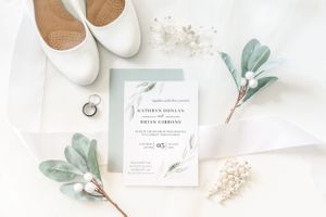 White wedding shoes, stationery, and greenery on a marble surface.