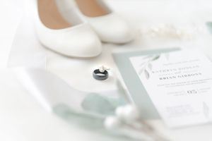 Close-up of white wedding shoes and stationery on a marble surface with greenery.