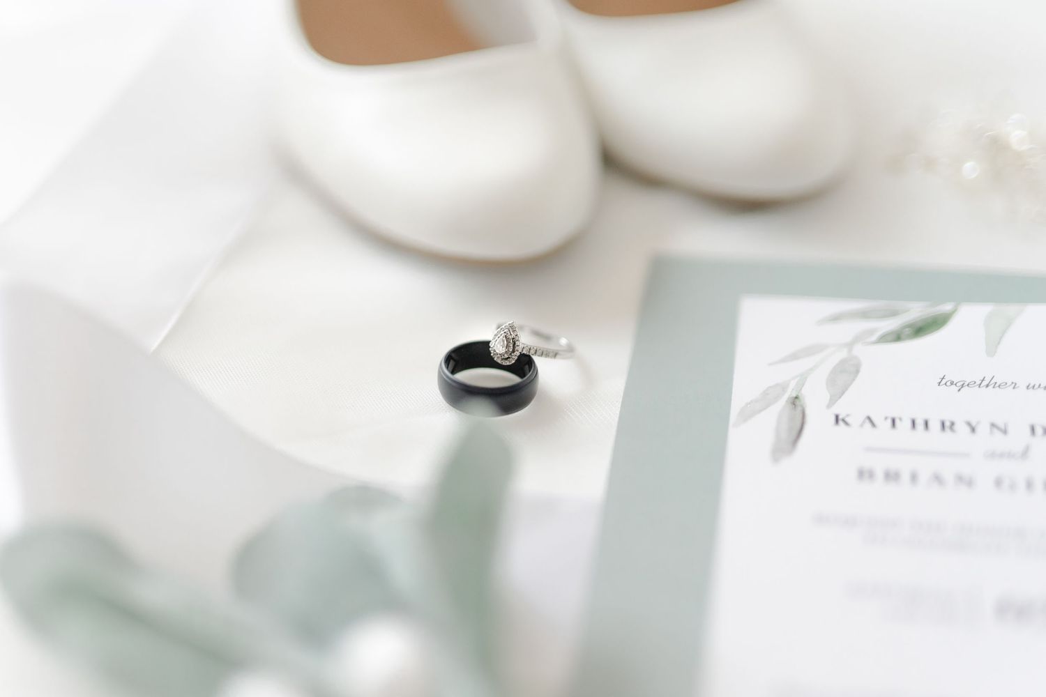 Close-up of white wedding shoes, stationery, and greenery on a marble surface.