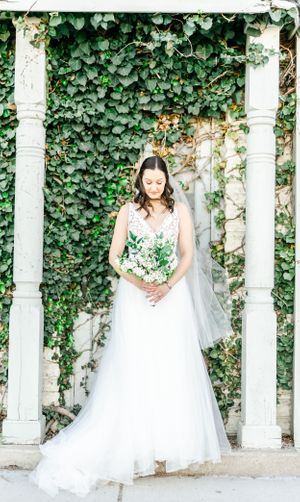 A woman in a white wedding dress stands in a lush, green garden surrounded by ivy-covered walls.