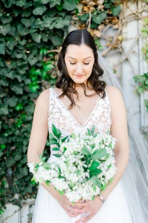 A woman in a white wedding dress holds a bouquet of flowers and stands in a garden with ivy-covered walls.