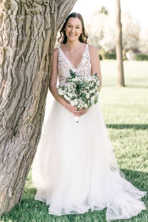 A woman in a white wedding dress stands in a grassy field, holding a bouquet of flowers and leaning against a tree.