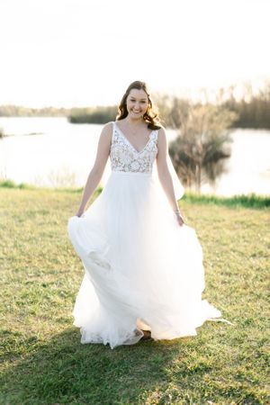 A woman in a white wedding dress stands in a grassy field, holding a bouquet of flowers and smiling.