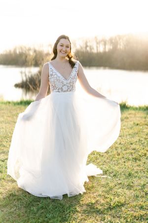 A woman in a white wedding dress stands in a grassy field, holding a bouquet of flowers and smiling.
