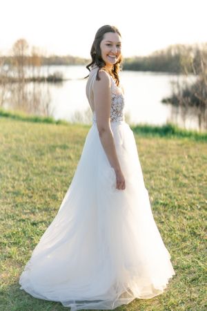 A woman in a white wedding dress stands in a grassy field, holding a bouquet of flowers and smiling.