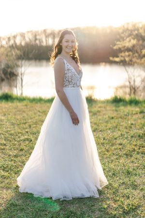 A woman in a white wedding dress stands in a grassy field, holding a bouquet of flowers and smiling.