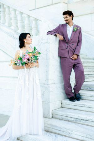 A couple in wedding attire stand on steps, with the woman holding a bouquet of flowers and the man in a purple suit.