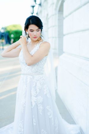 A woman in a white wedding dress stands on steps, with one hand on her face and a thoughtful expression.