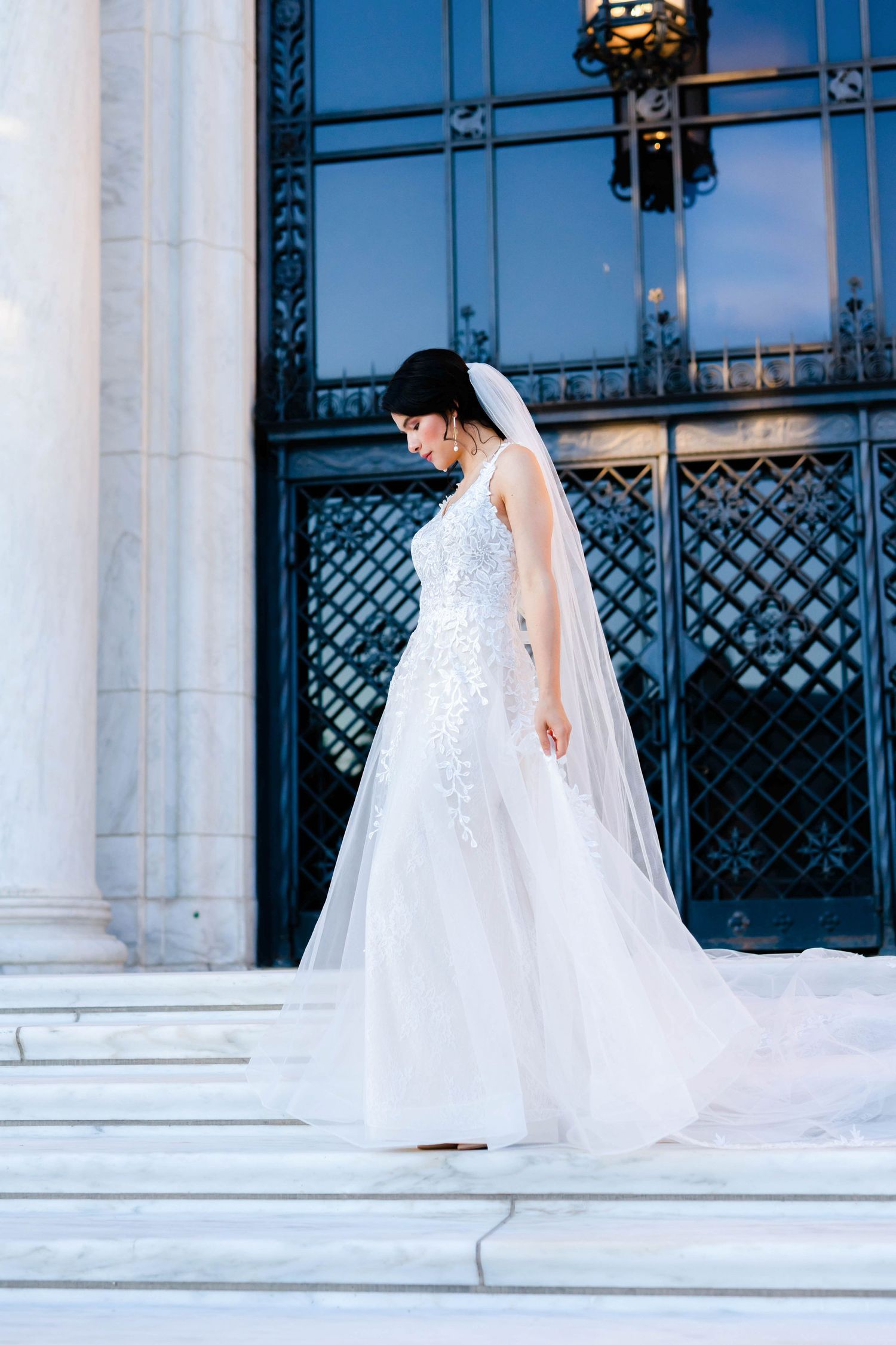 A woman in a white wedding dress stands on steps, holding a bouquet of flowers.