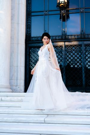 A woman in a white wedding dress stands on steps, holding a bouquet of flowers.