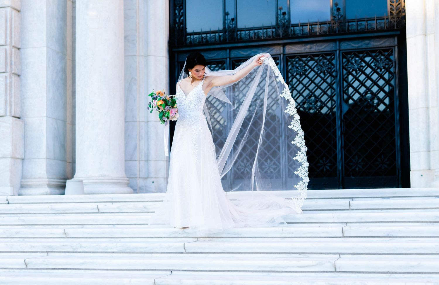 A woman in a white wedding dress stands on steps, holding a bouquet of flowers and looking up.
