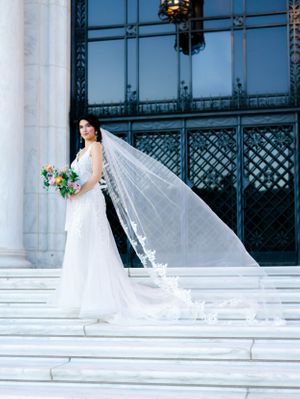 A woman in a white wedding dress stands on steps, holding a bouquet of flowers and looking at the camera.