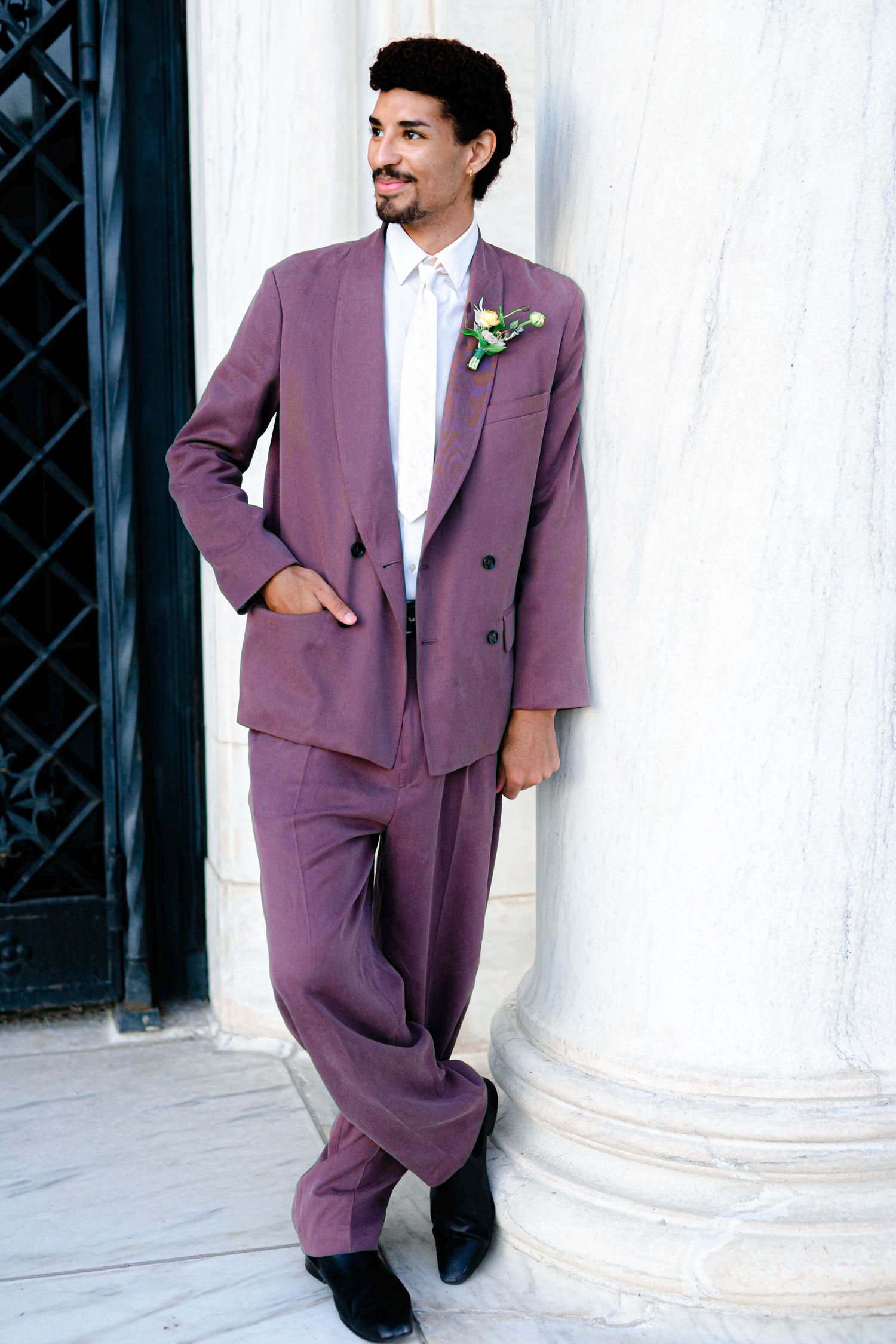The image shows a man wearing a purple suit and standing in front of a white column.