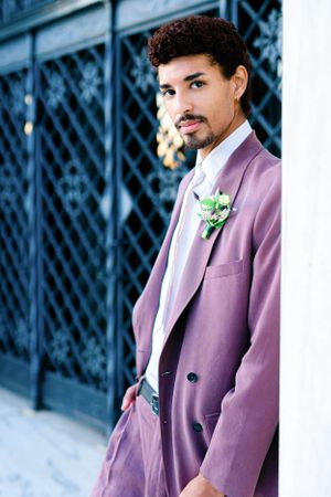 The image shows a man wearing a purple suit and standing in front of a white column.