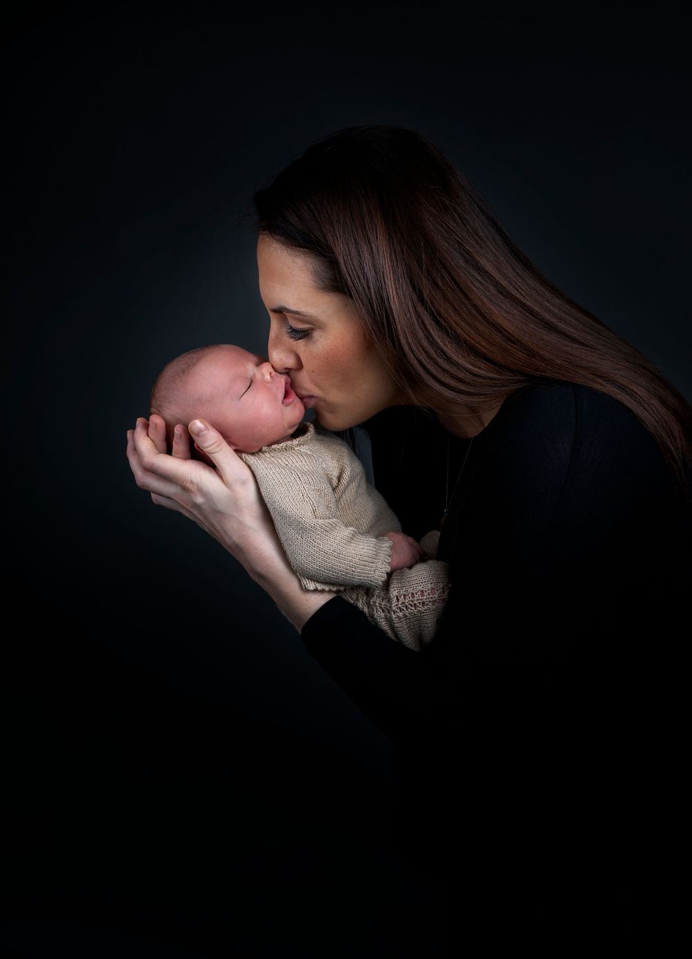 Dramatic low-key portrait of parent kissing newborn baby wrapped in neutral tones.