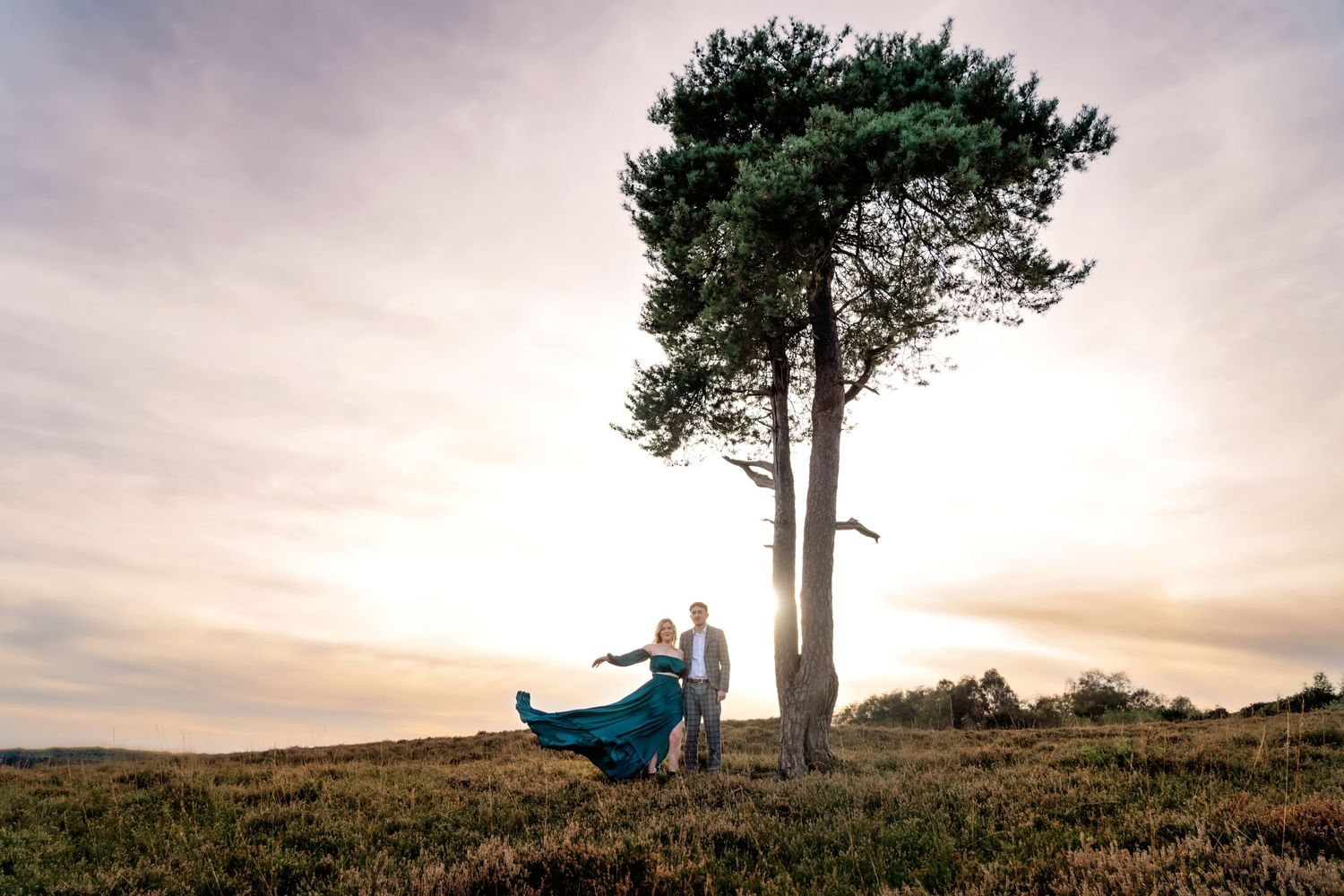 Engagement photoshoot in the New Forest with a beautiful tree and sunset sky in the background, with the gown flying in the wind.
