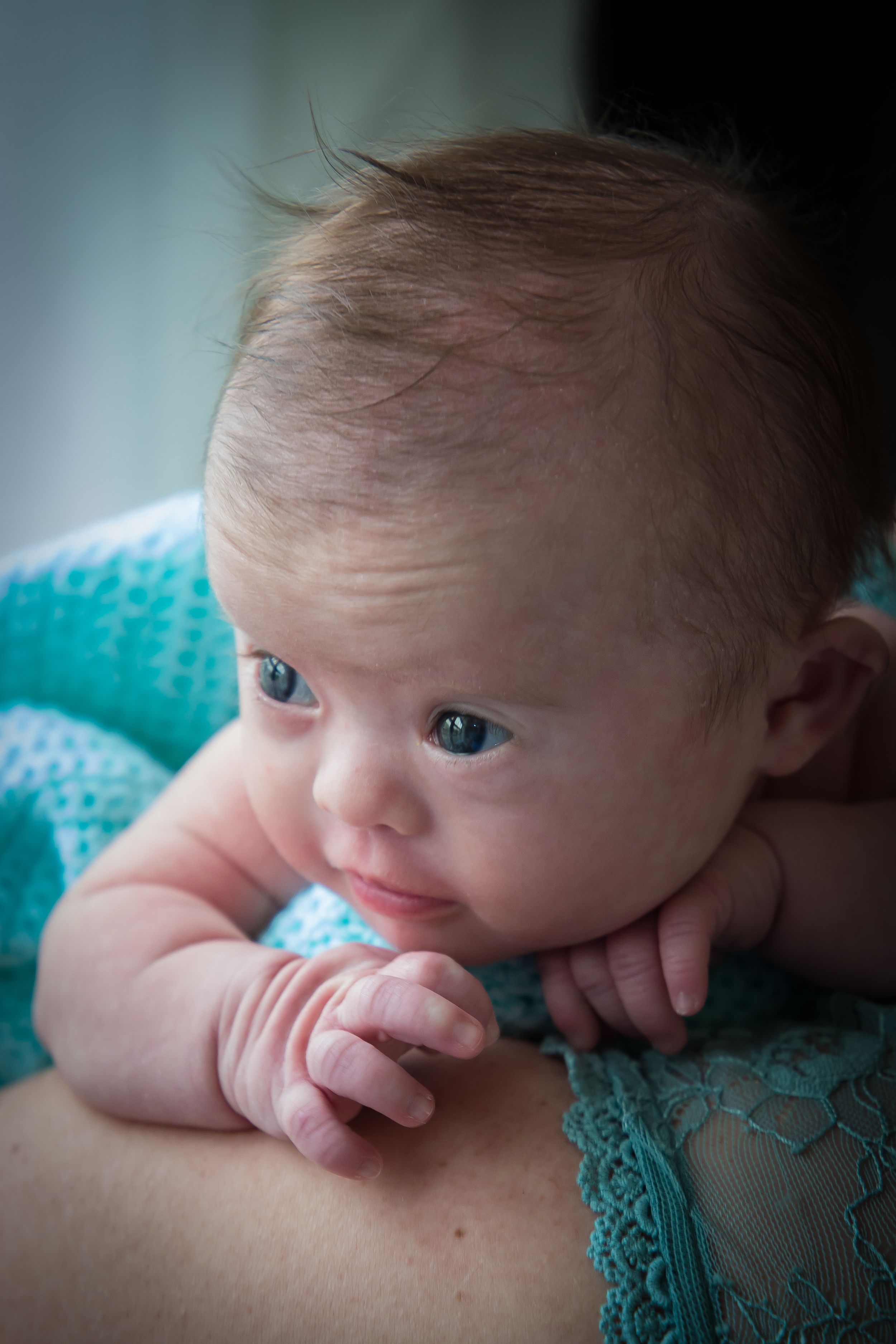 A baby with bright eyes rests on a turquoise knit fabric in an intimate portrait.