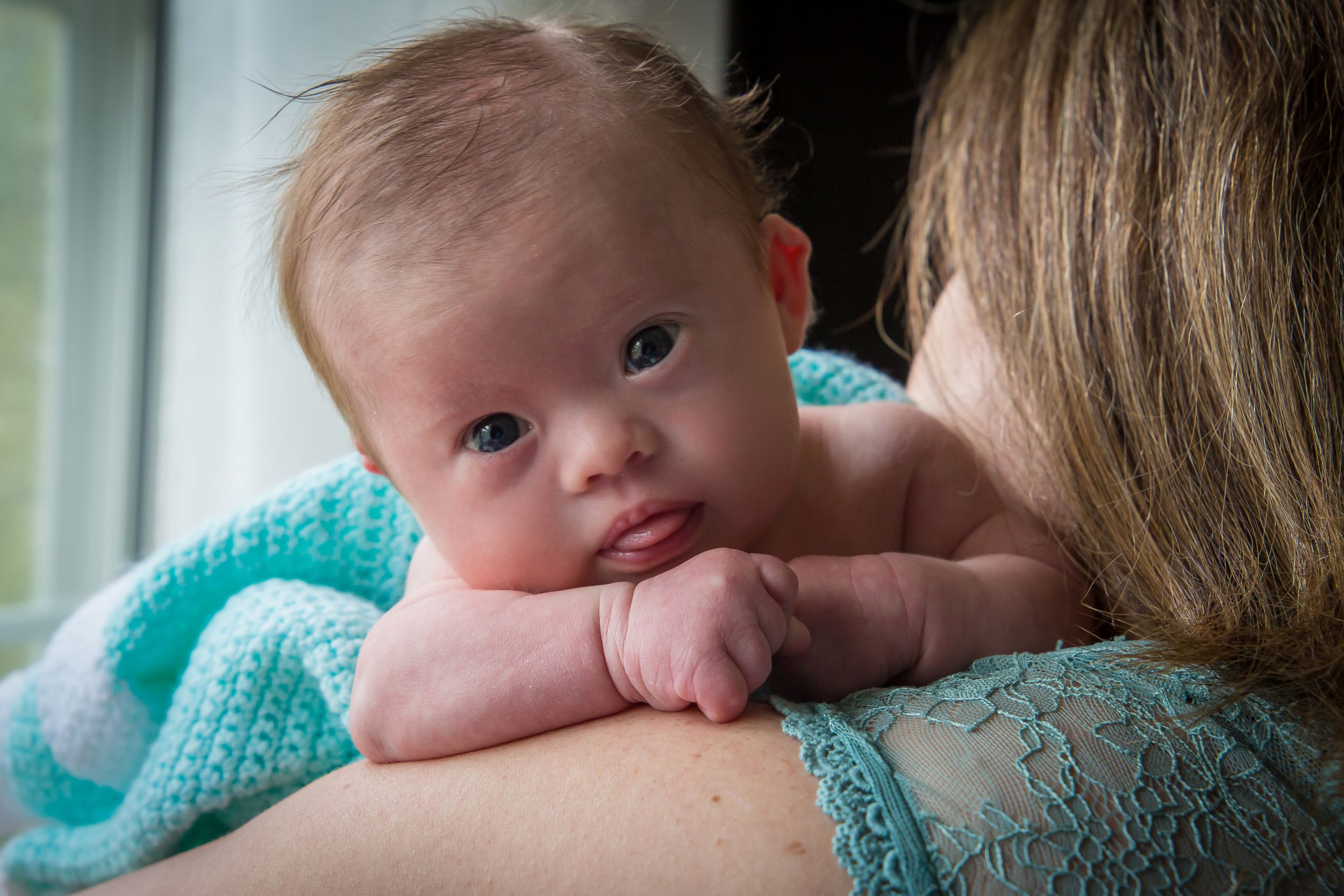 A baby rests against someone wearing a turquoise knit sweater in soft lighting.