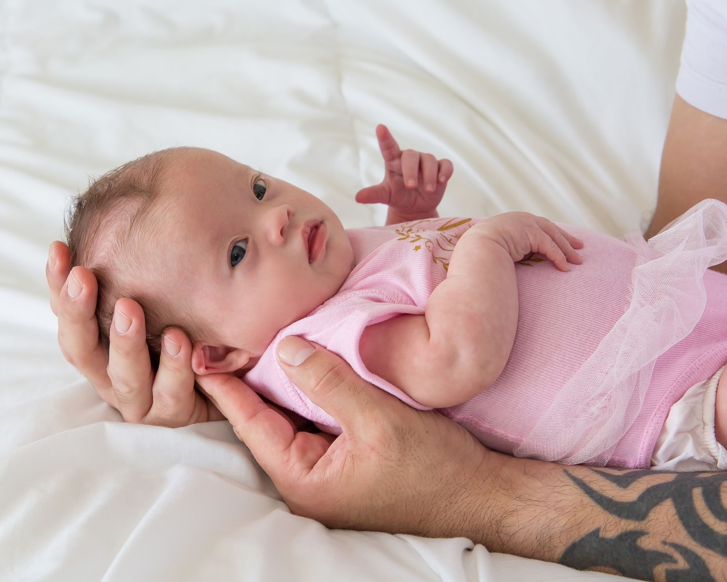 A newborn baby rests on white bedding while being gently supported by hands.