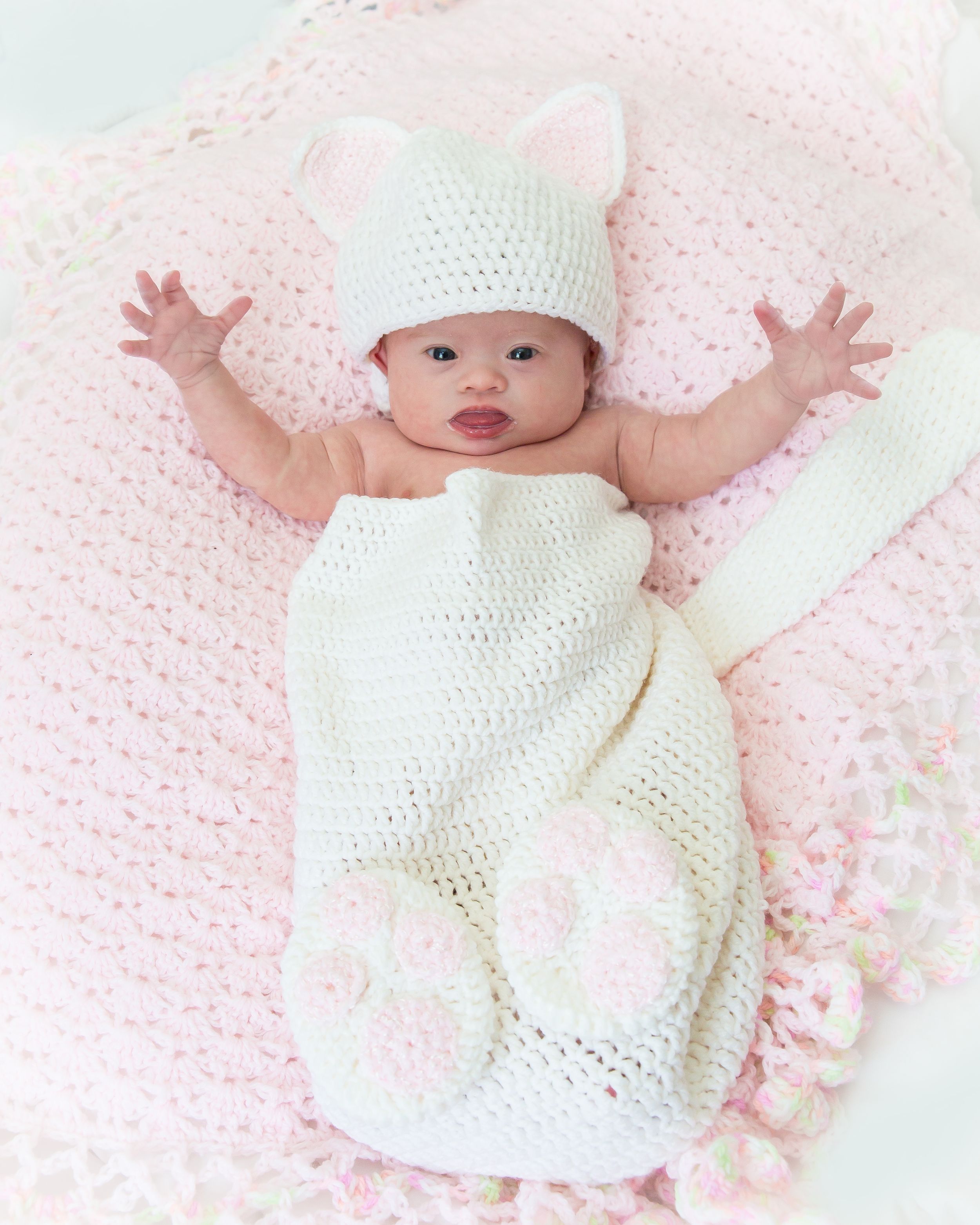 A baby wearing a white knit outfit and hat lies on a light pink background.