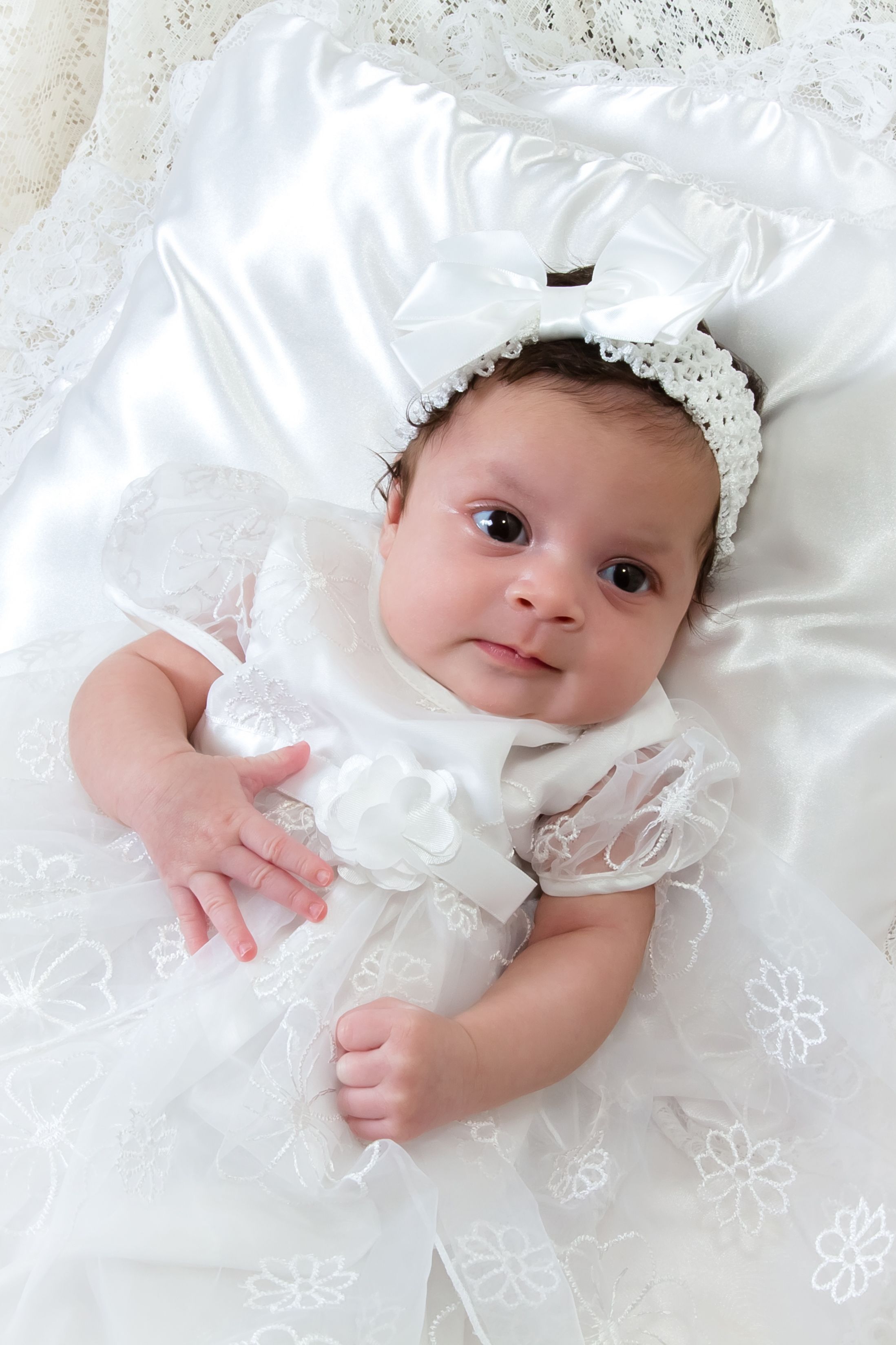 Baby in white christening dress and bonnet lying on lace bedding.
