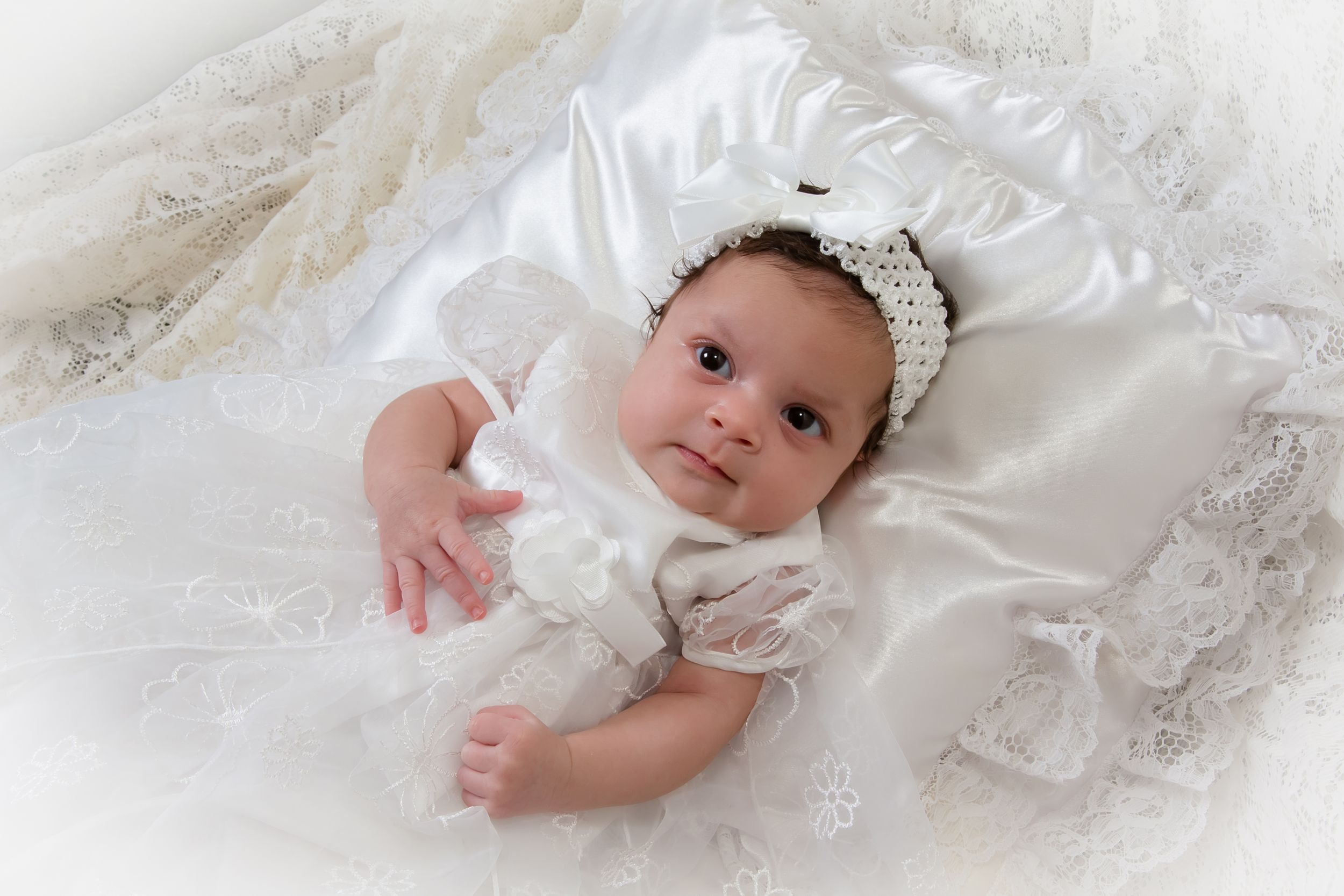 Baby in white christening dress posed on white lace backdrop.
