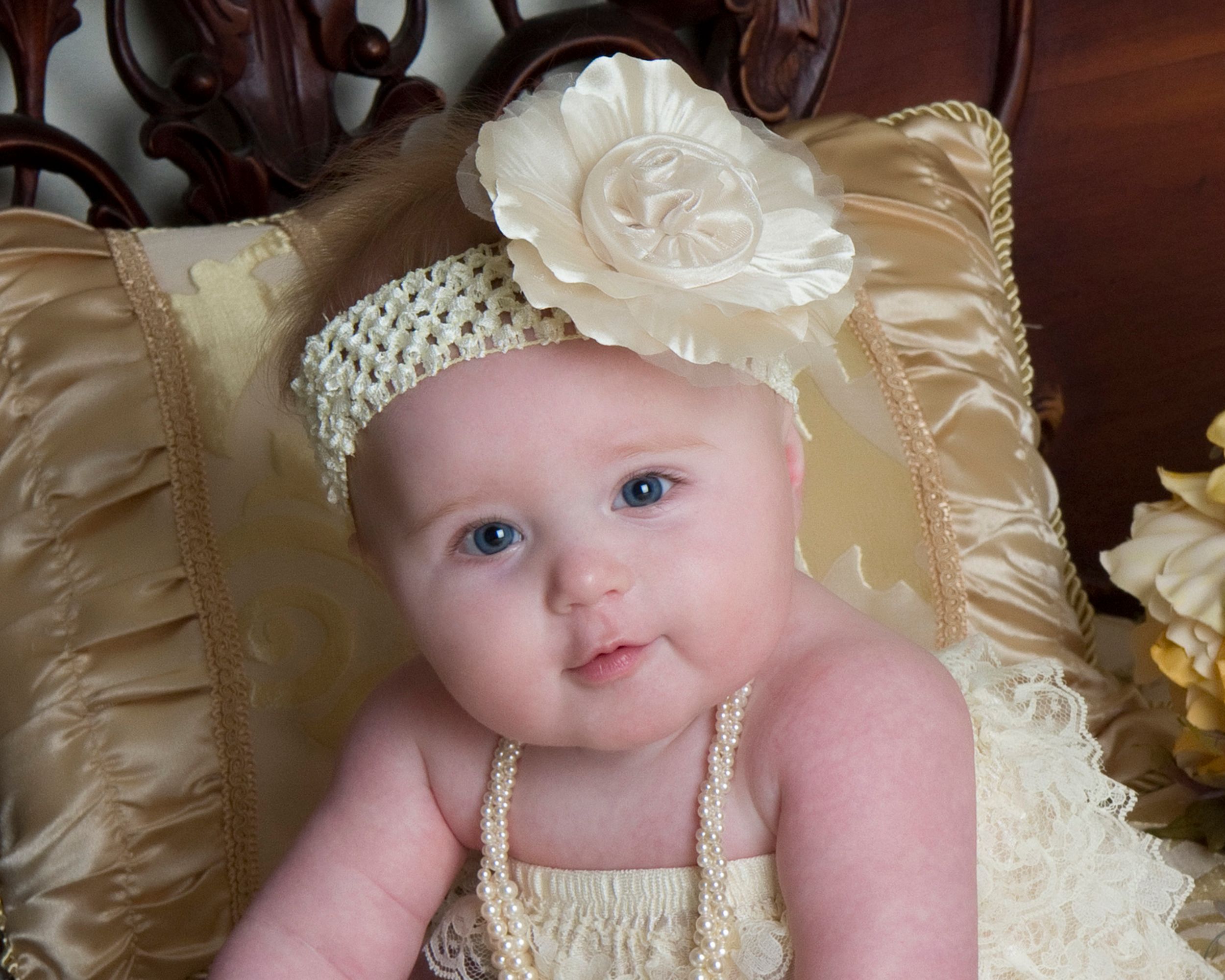 Baby wearing pearl headband and cream dress posed on silk pillows.