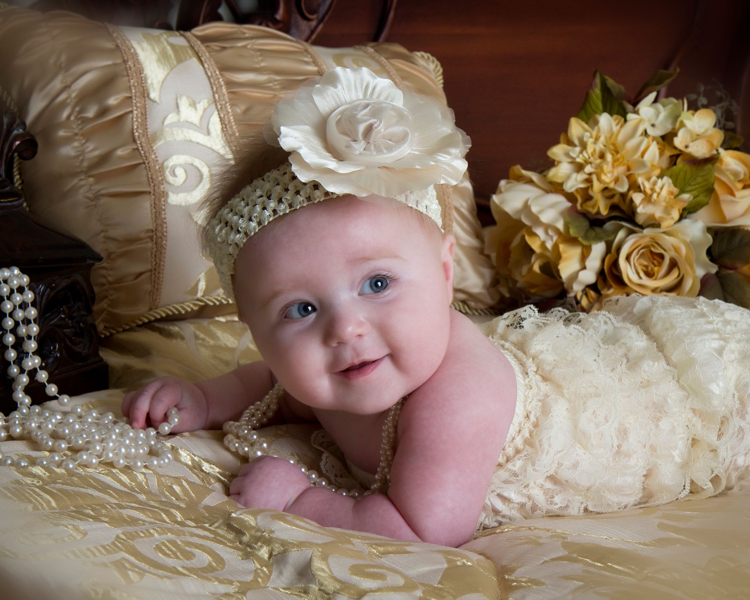 Baby in cream dress with floral headband posed on golden pillows.