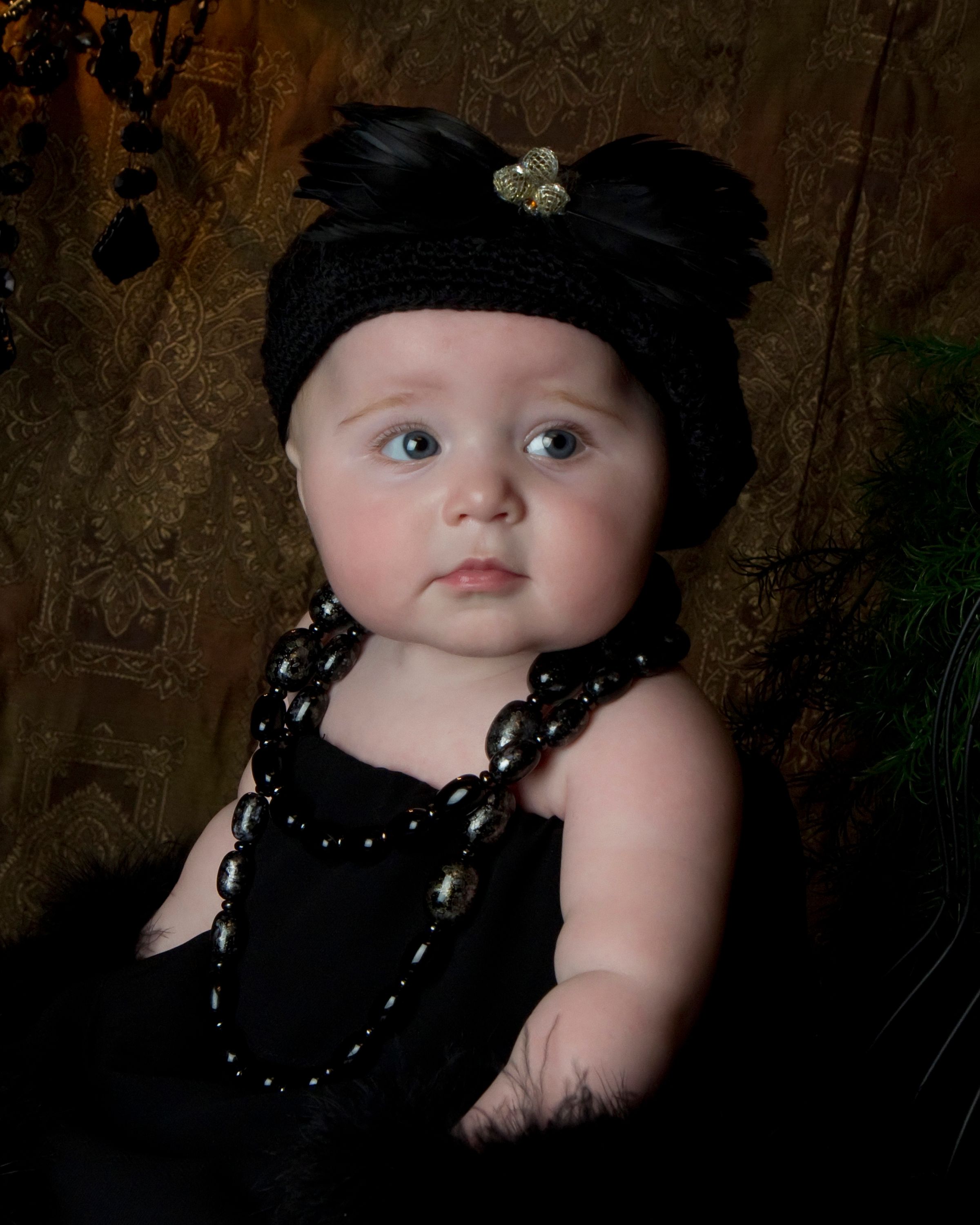 Baby in black outfit with beaded necklace against dark background.
