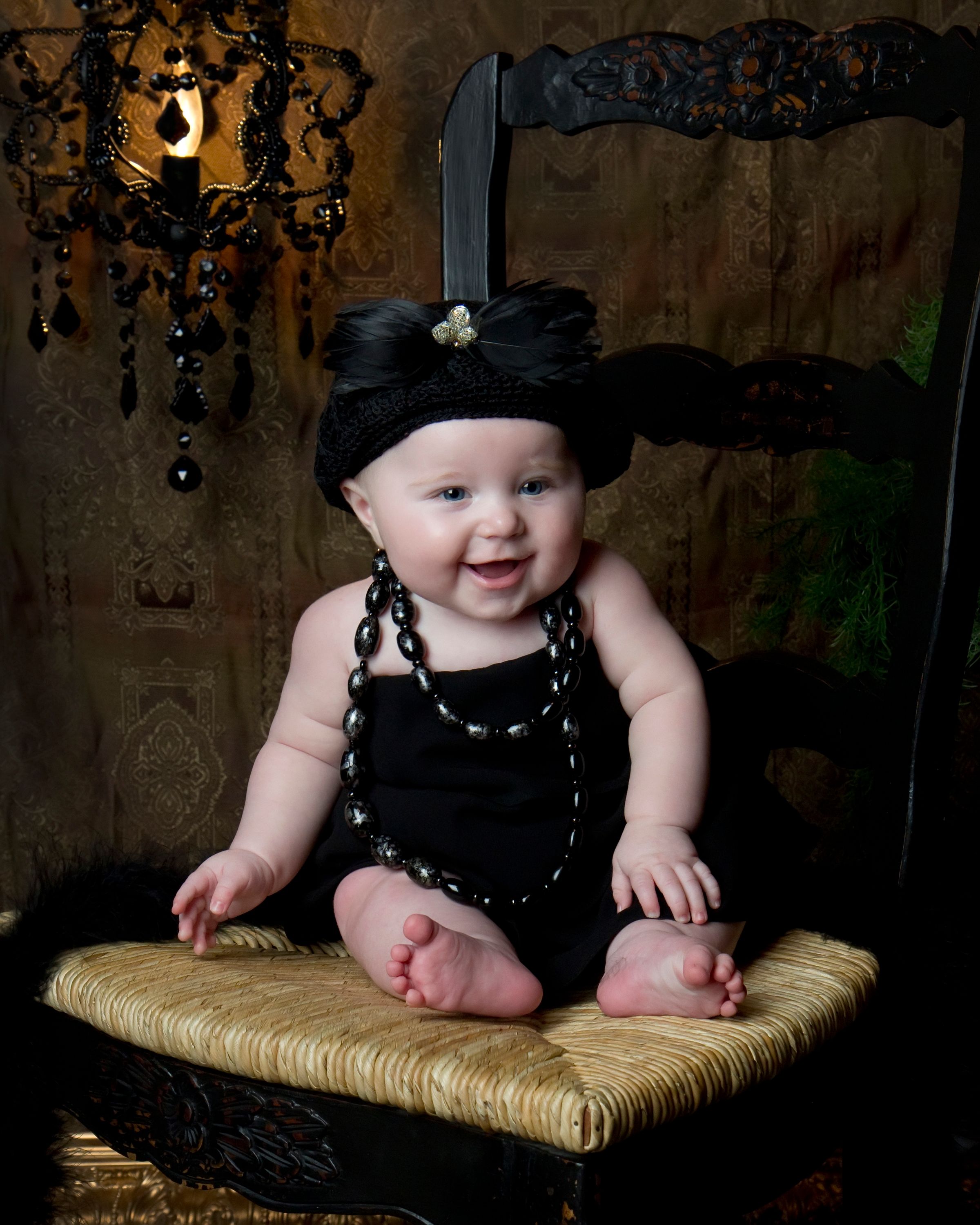 A small baby wearing a black dress and hat sits on a woven chair against a dark background.