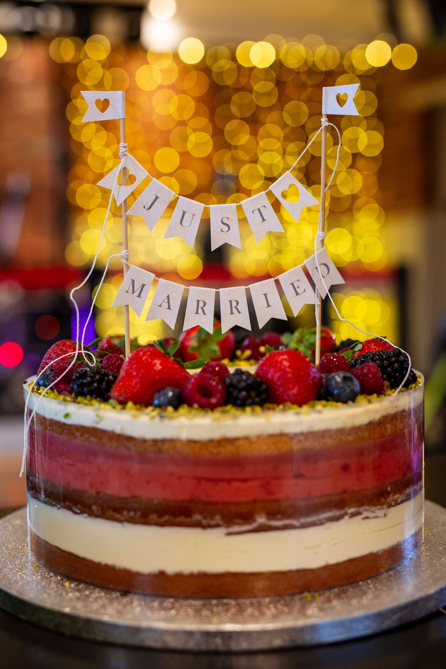 Wedding cake decorated with fresh berries and Just Married bunting against golden bokeh lights.