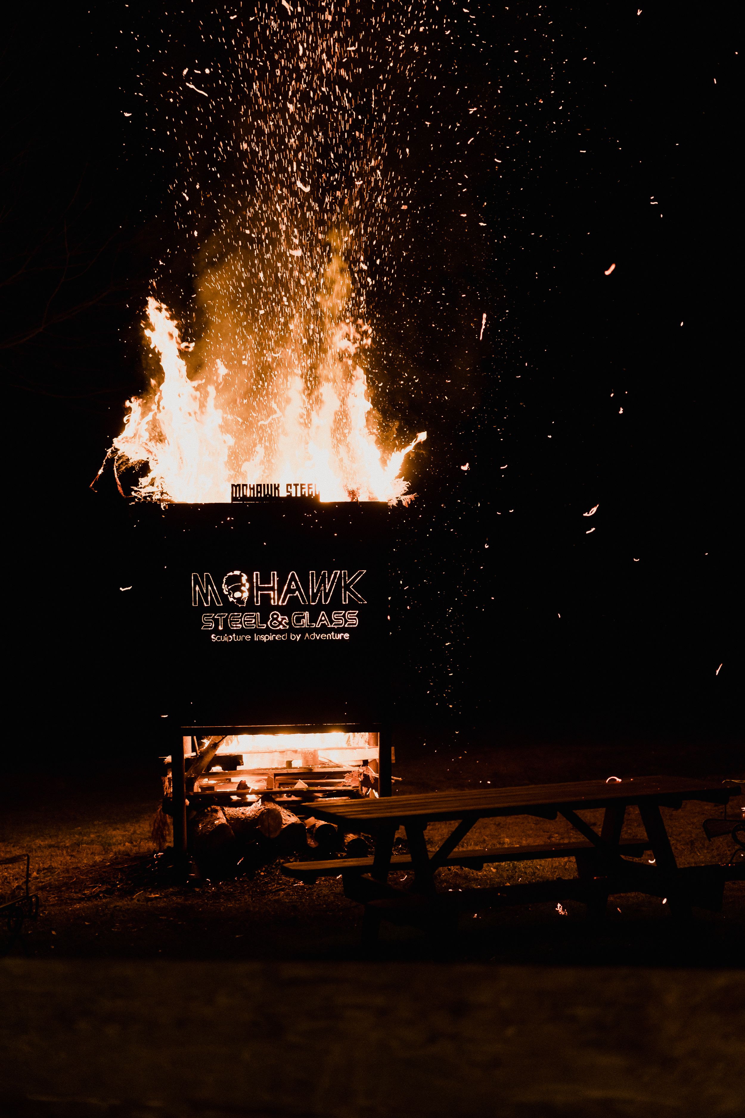 Large bonfire sending sparks into the night sky with text illuminated in the foreground.