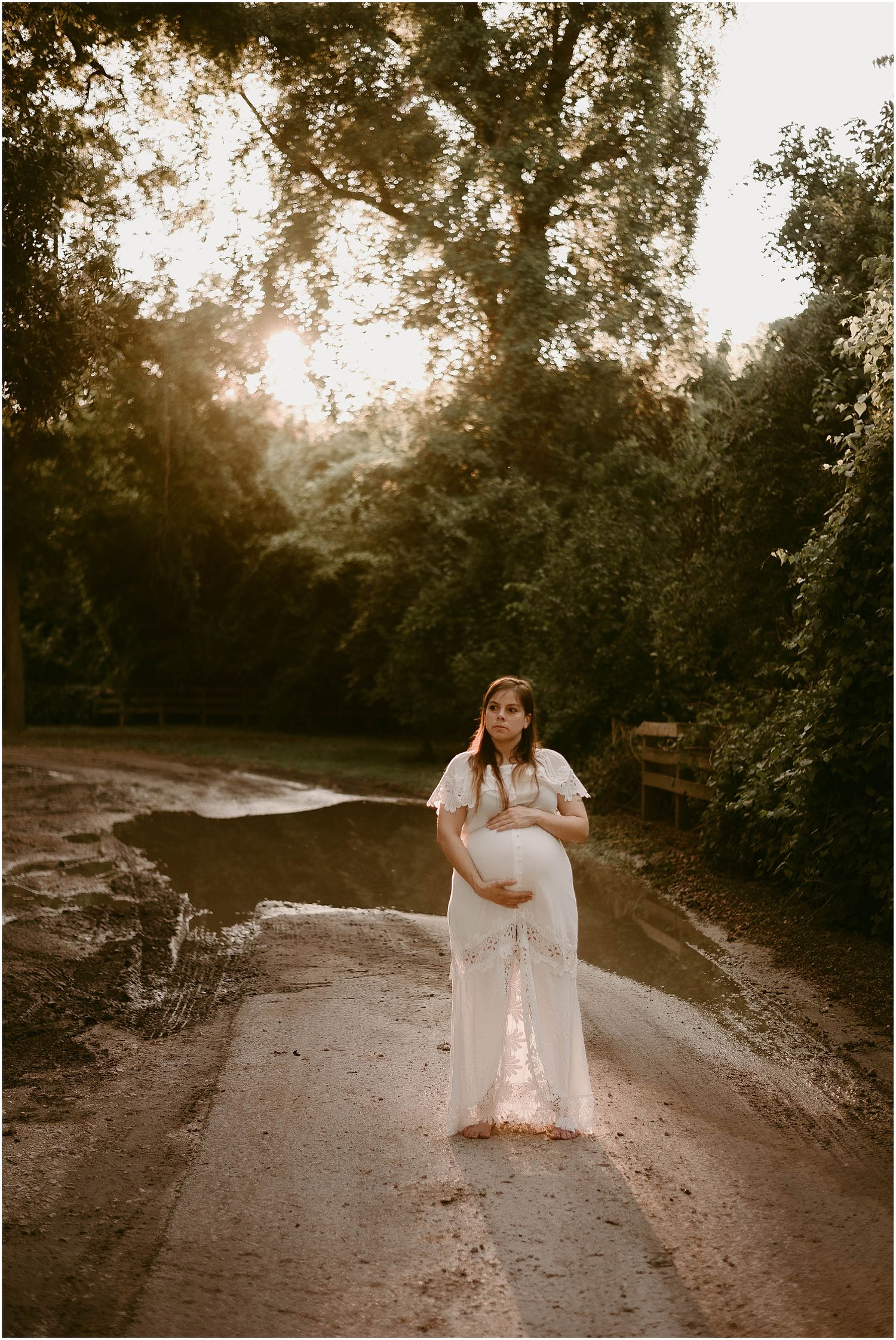 Full-length maternity portrait with sunlit trees creating dreamy backdrop on dirt path.