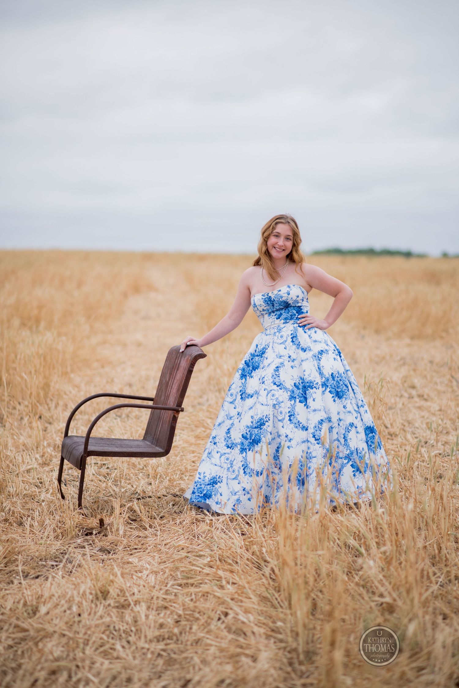 Fashion portrait with vintage chair prop showcasing blue floral ballgown in rural field setting.