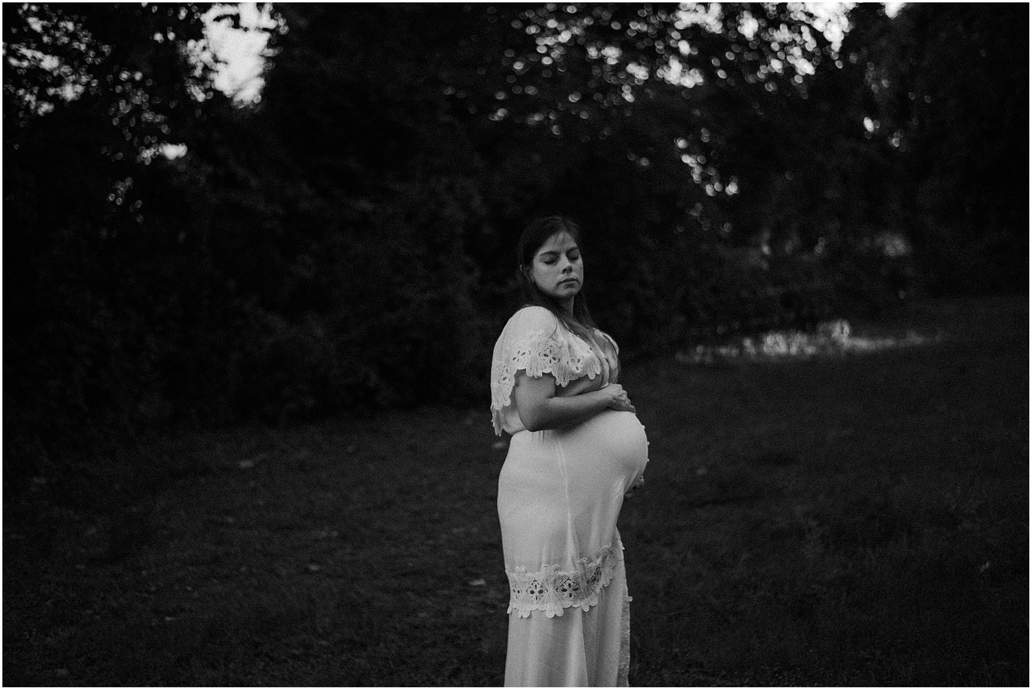 Black and white maternity photo showing an expectant mother in a flowing white dress outdoors.