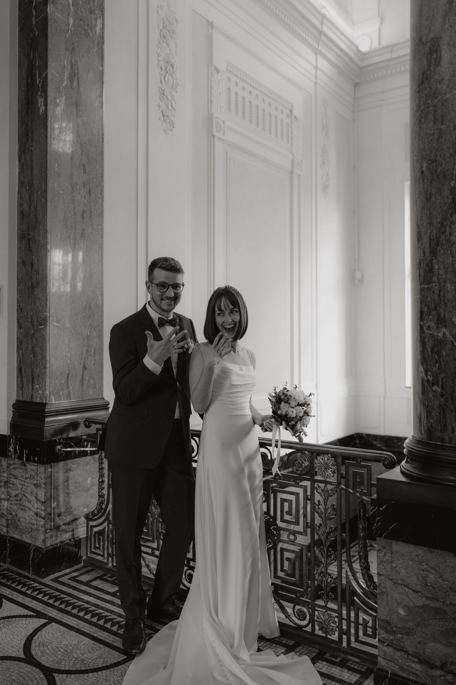 Black and white photo of elegant couple in formal attire standing in ornate marble room.