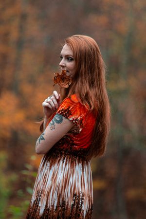 A person with long red hair in vintage dress holding autumn leaves against blurred fall forest background.