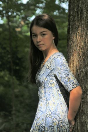 Someone wearing a light blue patterned dress poses against a tree trunk in a forest setting.